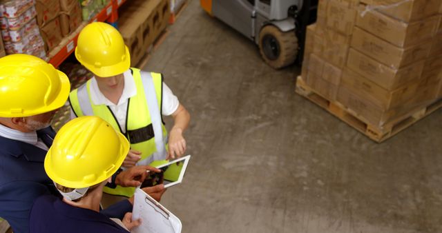 Workers in warehouse wearing high-visibility gear and helmets planning logistics together. They are using tablet and clipboard for inventory management and stock organization. Ideal for content about supply chain, safety standards, team collaboration, and warehouse operations.