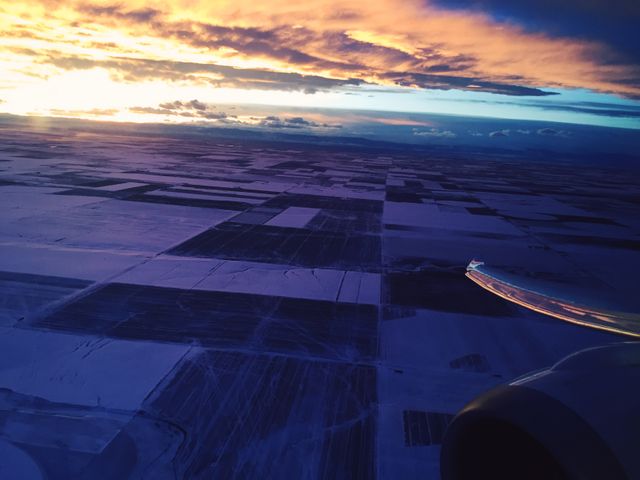 Aerial view capturing snow-covered farmland fields during a vibrant winter sunset, seen from an airplane. The colors of the setting sun meld with the patchwork of fields below, creating a tranquil and scenic landscape. Ideal for travel brochures, winter-themed content, and nature photography collections showcasing the serene beauty of agricultural land from above.