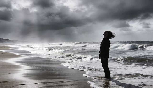 Solitary man standing on windswept beach with dramatic storm clouds and crashing waves