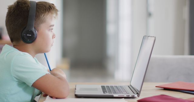 A young boy is sitting at a desk using a laptop with headphones on, likely engaged in online learning or educational activities. The image can be used for educational content, remote learning resources, e-learning platforms, and articles about modern education and technology in use at home.