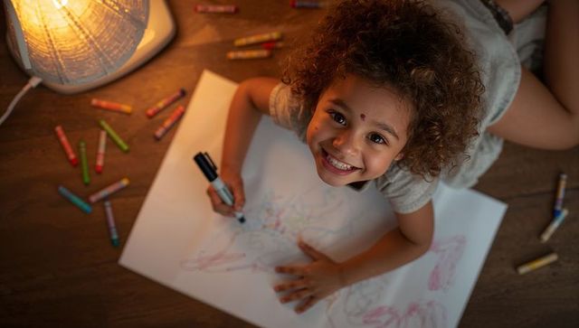 Smiling child drawing on floor by warm lamp holding marker with crayons scattered