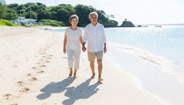 Senior couple walking hand in hand on sunlit tropical beach barefoot with footprints