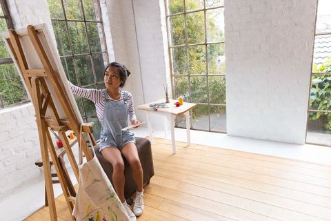 Chinese woman painting on easel in sunlit studio wearing denim overalls and striped top, natural lig