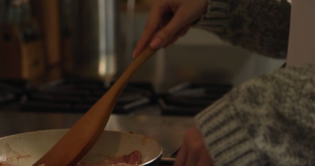 A close-up detail of a person using a wooden spoon to cook bacon in a skillet on a stovetop, perfect for food blogs, cooking websites, recipe illustrations, and culinary articles that focus on home cooking, kitchen appliances, and utensils. Ideal for social media content, culinary education, and promotions for cooking equipment.