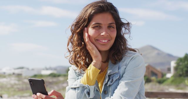 Portrait of happy biracial woman holding smartphone and smiling on sunny promenade by the sea. leisure time, outdoors and close to nature.