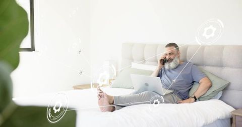 Bearded senior working remotely on bed with laptop and phone, cozy home office lifestyle