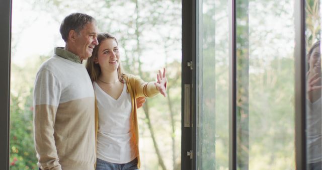 Smiling adult daughter with arm around senior father standing and looking outside window at home. Useful for concepts of family bonding, parent-child relationships, and happiness within a home setting. Suitable for articles, blog posts, and advertisements focusing on family life, caregiving for elderly parents, and intergenerational connections.