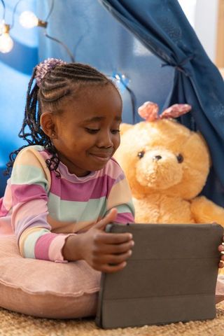 African american girl using tablet in cozy play tent with teddy bear and string lights
