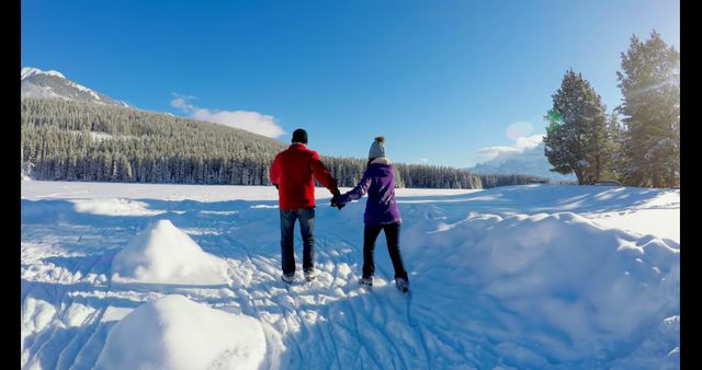 Couple is walking hand in hand through a snowy landscape with a view of mountains in the background under a clear, sunny blue sky. Ideal image for themes related to winter vacations, romantic getaways, outdoor adventures, nature exploration, and seasonal travel promotions.