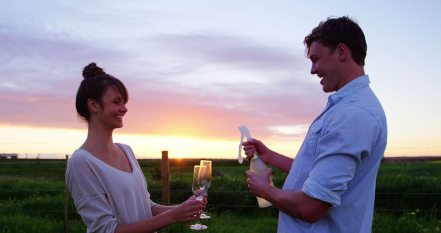 Couple celebrating outdoors with champagne at sunset. They are smiling and holding champagne glasses while in a green field during sunset. Suitable for use in concepts related to romance, celebration, special occasions, love, and happiness.