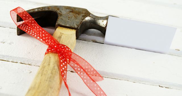 A hammer with a wooden handle is wrapped in a neat red ribbon with white polka dots, resting on a white wooden surface. The festive combination suggests the tool could be a gift or part of a celebration. Suitable for use in online articles, blogs, or advertisements related to DIY projects, holidays, and gifting ideas.