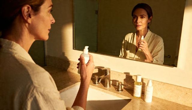 Sunlit golden hour skincare ritual with mature woman holding pump bottle at bathroom vanity