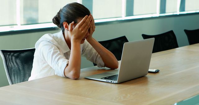 A young Caucasian woman appears stressed or frustrated at her workplace, with her hands covering her face as she sits in front of a laptop. Her body language suggests she might be dealing with a challenging situation or feeling overwhelmed by work.