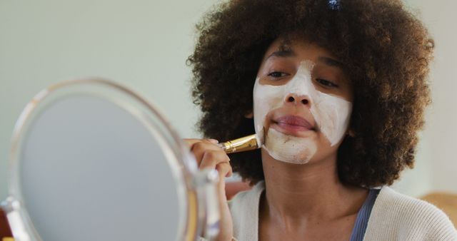 Happy biracial woman applying beauty face mask with brush. spending quality time at home alone.