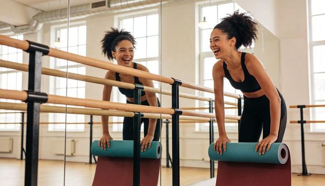 Smiling woman leaning on ballet barre holding rolled exercise mat in sunlit dance studio