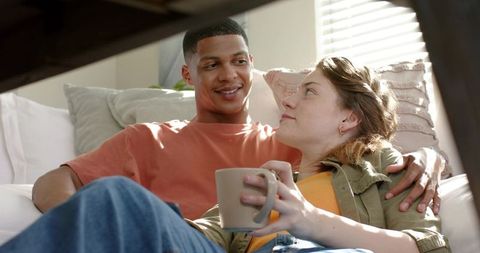 Multiracial couple lounging on sunlit sofa sharing coffee and intimate moment
