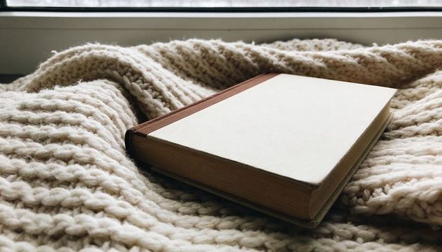 Resting hardcover journal on chunky knit blanket by window in soft daylight cozy reading nook