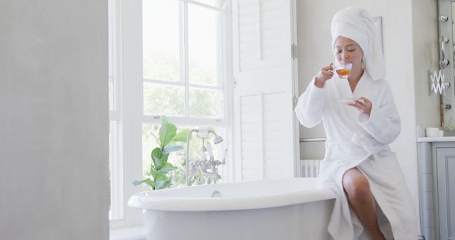 Shows a woman enjoying a relaxing moment in her bathroom while drinking tea. Ideal for topics related to wellness, self-care routines, morning rituals, and creating tranquil spaces at home. Could be used in advertisements for bath products, teas, spas, or wellness blogs.