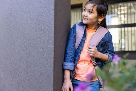 Young asian girl leaning by column holding pink backpack looking right with bokeh