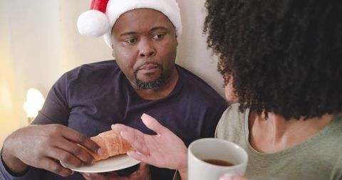 African American Couple Wearing Santa Hat Sharing Coffee and Croissant, Conversing Warmly