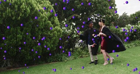 Two graduates walking on lawn in caps and gowns holding diplomas amid purple confetti