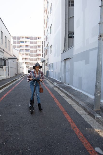This image depicts a biracial woman with short blonde hair, wearing a hat and denim dungarees, riding an electric scooter in a city alley. The scene is set on a sunny day, highlighting urban lifestyle and modern transportation. Ideal for use in articles or advertisements focusing on urban living, independent women, eco-friendly transportation, or modern commuting solutions.