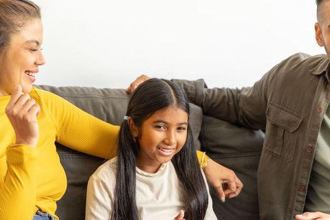 Smiling girl sitting between parents on cozy sofa, multicultural family sharing warm moment
