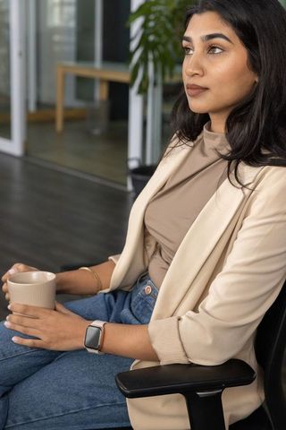Indian businesswoman sitting in office lounge holding coffee mug, looking contemplative