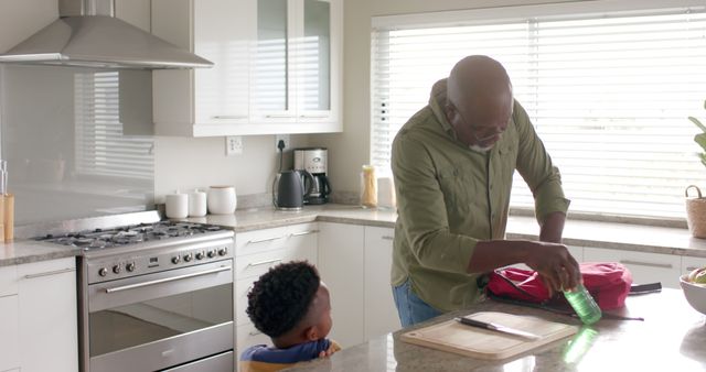 Grandfather preparing lunch for his grandson in a bright, modern kitchen. Man packing lunch container while young boy watches attentively. Ideal for depicting family values, multi-generational bonding, home life, and everyday routines. Excellent for advertising family-oriented products or services, or illustrating lifestyle articles.