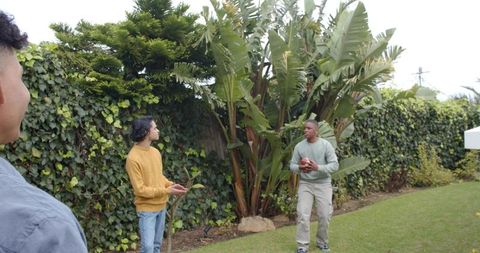 Friends tossing football on backyard lawn near lush hedge and tropical plants casual outdoor play