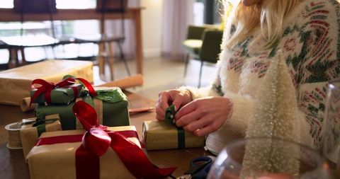 Woman wrapping holiday gifts with kraft paper and satin ribbons on cozy sunlit table