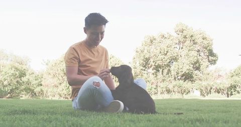 Asian man sitting cross-legged feeding black dog in park enjoying relaxed companionship