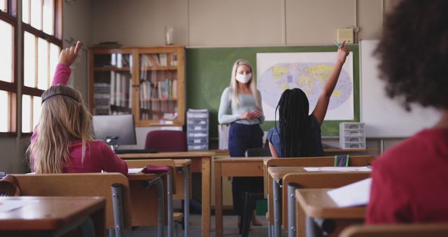 Teacher wearing a mask interacts with engaged students in classroom. Ideal for educational content, classroom management resources, pandemic relation education materials, and interactive learning promotion.