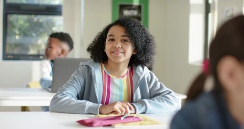 Smiling student sitting at desk in bright classroom wearing gray hoodie and colorful stripes