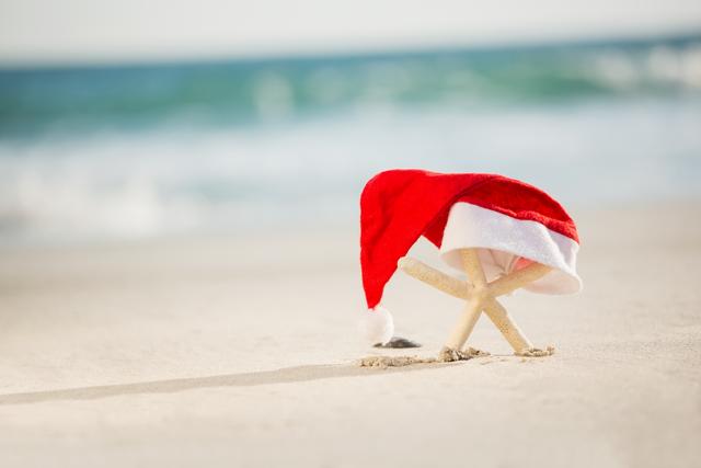 Starfish wearing Santa hat on beach sand with ocean in background. Perfect for holiday-themed travel promotions, Christmas vacation advertisements, festive greeting cards, and tropical holiday decor.
