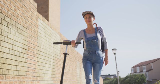 Biracial woman wearing cap and overalls walking with scooter on street - Download Free Stock Photos Pikwizard.com