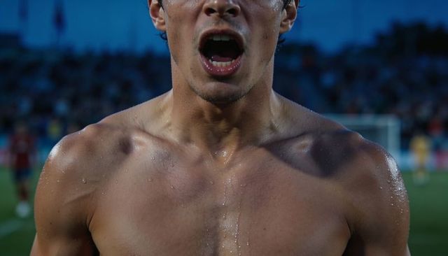 Shouting shirtless soccer player sprinting on pitch at evening match, sweat and intensity