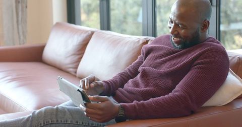 African american man relaxing on leather sofa using tablet at home, modern lifestyle