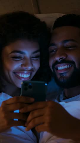 Couple cuddling in bed watching phone, laughing and bonding | Vertical night video