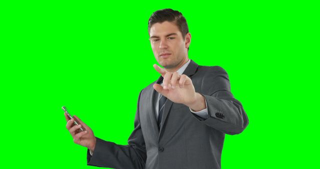 A young Caucasian businessman in a suit gestures towards the camera while holding a smartphone, with copy space on the green background. His confident expression and hand gesture suggest he's presenting or interacting with a digital interface.