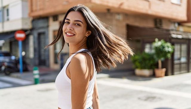 Smiling woman turning hair while walking on sunny urban street in white crop top