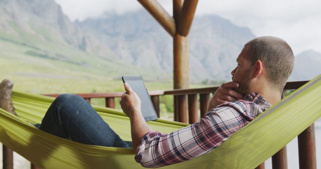 Man relaxing in green hammock using a tablet in a mountainous area with scenic views. Ideal for concepts related to relaxation, technology, and the enjoyment of nature. This can be used in travel brochures, websites promoting tranquil getaways, technology ads, and lifestyle blogs emphasizing work-life balance.