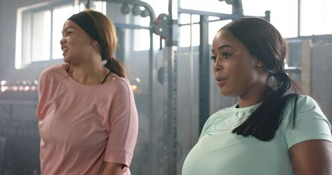 Diverse women catching breath and smiling after strength workout in gym, pastel activewear