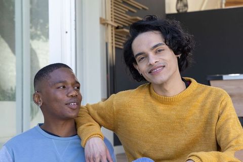 Young men sitting on sunlit porch sharing warm moment, smiling in casual knit sweaters