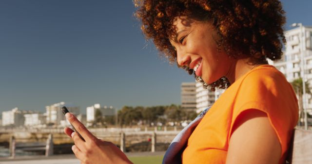 Woman with curly hair texting on smartphone. Attractive woman in casual attire enjoys sunny day outside city buildings. Great for themes of modern communication, living in urban areas, and outdoor activities.