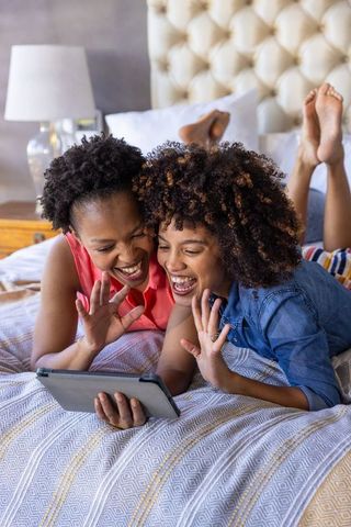 Laughing diverse female friends video chatting on tablet while lying on cozy bed