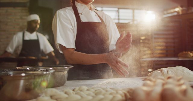 Baker preparing dough on a flour-dusted counter in a professional bakery kitchen. Includes another staff member in the background. Useful for advertisements, culinary articles, and food industry features highlighting professional baking, teamwork, or culinary skills.