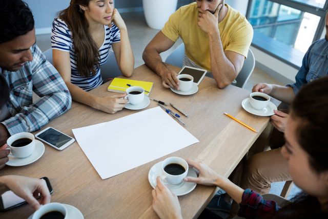 Group of young professionals collaborating on a project over coffee in a modern office. Ideal for illustrating teamwork, idea generation, startup culture, and collaborative work environments. Suitable for marketing materials, blog posts about creativity in the workplace, and business presentations.