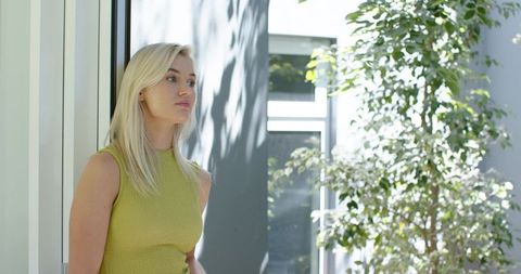 Gazing woman standing by glass door in modern atrium, sunlight casting leafy shadows