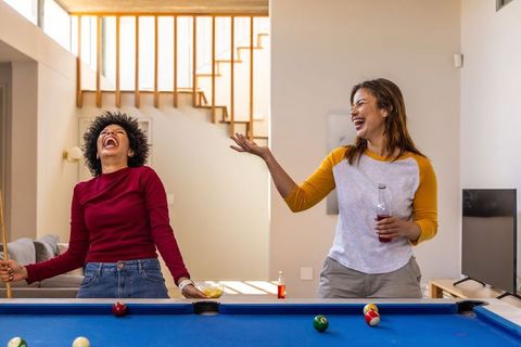 Diverse friends laughing and playing pool in sunlit modern living room holding cue and drink